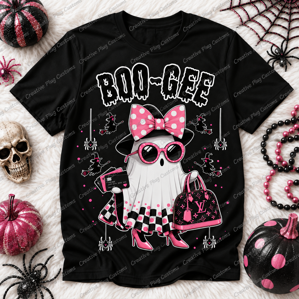 A black short-sleeve graphic T-shirt laid flat on a white faux-fur surface with Halloween props around it. The shirt features a ghost character wearing a pink polka-dot bow, pink sunglasses, a pink-and-black checkered skirt, and holding a pink handbag, with the text 'BOO-GEE' above.
