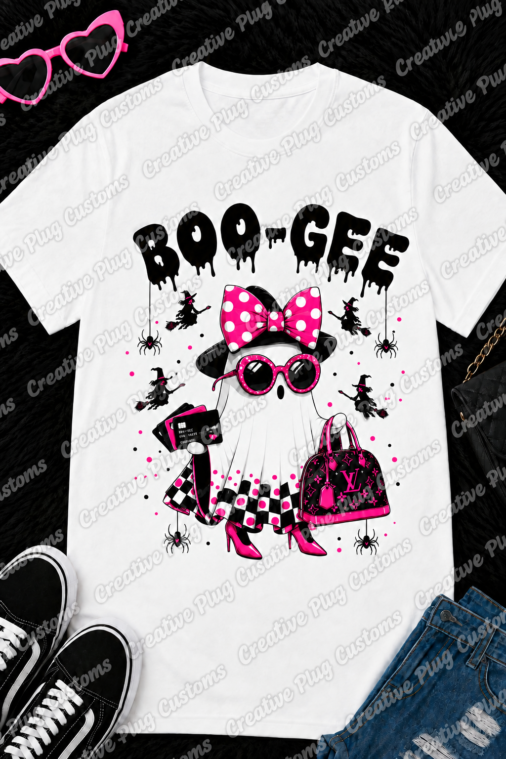 A white graphic t-shirt featuring a cartoon ghost dressed in pink and black Halloween attire with the text 'BOO-GEE' above it. The shirt is styled in a flat lay with jeans, sneakers, sunglasses, and a purse on a black textured background.