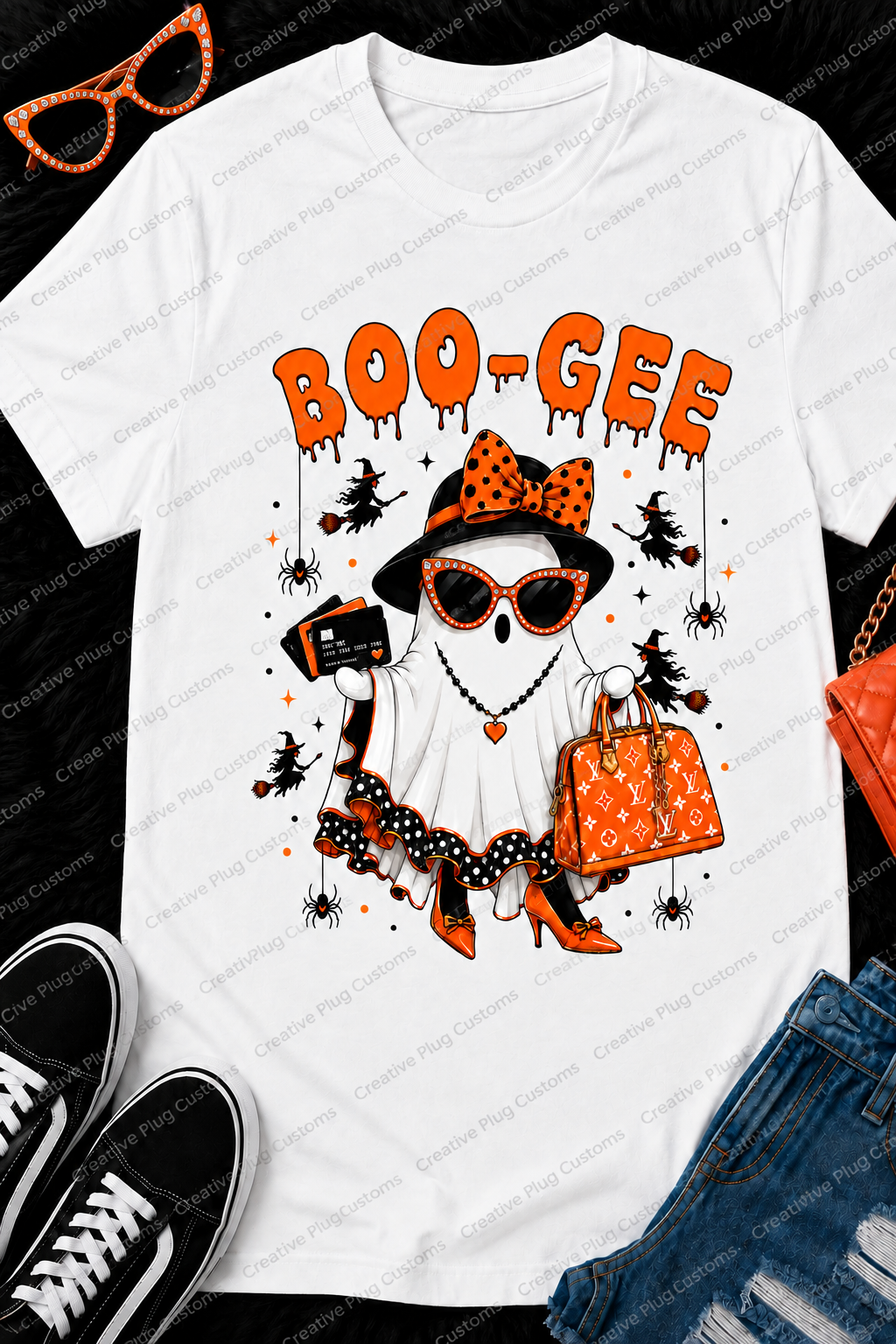 A white graphic t-shirt featuring a Halloween-themed ghost illustration with orange and black details, laid flat on a black textured background with styling props including sunglasses, shoes, jeans, and a quilted handbag.